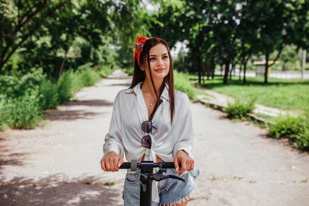 Linda chica morena monta en un scooter de electrodos en el parque que lleva una camisa blanca. concepto de transporte ecológico y alquiler.