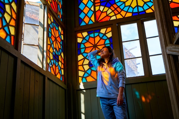 Linda chica morena posando en el auténtico balcón de un antiguo edificio residencial con una vidriera de mosaicos multicolores.