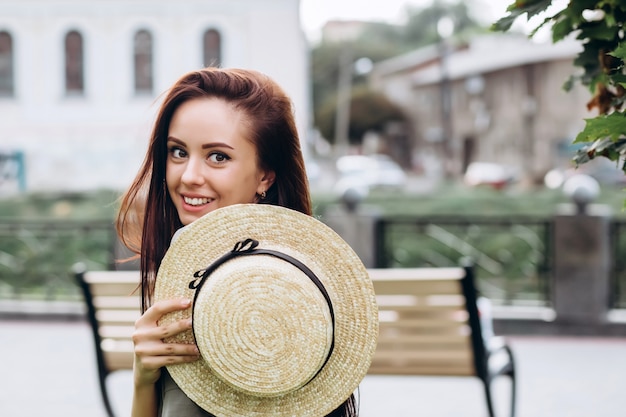 Linda chica morena con un sombrero de paja al aire libre, niña sonriente camina por la ciudad en el verano