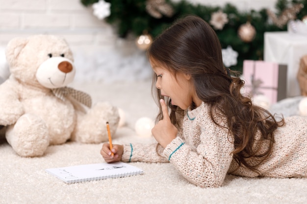 Linda chica con osito de peluche escribiendo una carta a Santa Claus en casa