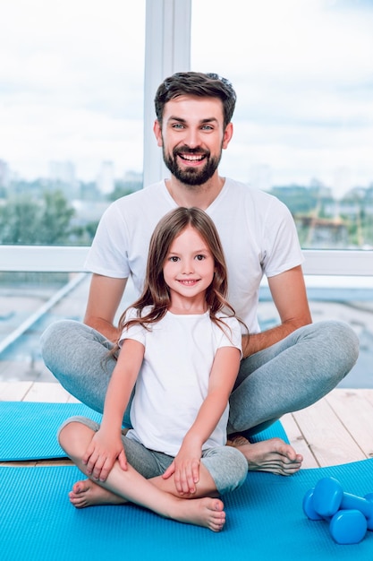 Linda chica y papá sentados en posición de loto en colchonetas de yoga junto a la ventana y sonriendo a la cámara