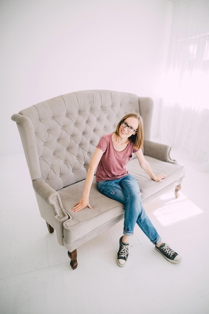 Linda chica pelirroja con camisa burdeos sentada en un sofá de cuero en una habitación luminosa con diseño de loft sonriendo