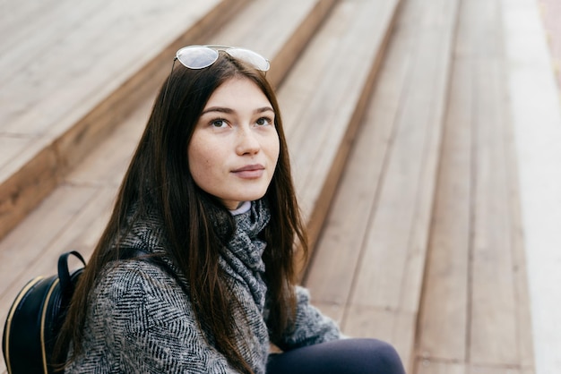 Linda chica de pelo oscuro en abrigo gris se sienta en pasos al aire libre, estilo callejero