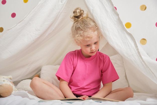 Una linda chica con el pelo rizado lee un libro en una cabaña de una sábana en la cama, una niña divirtiéndose y jugando en su tienda de campaña, Hut en la habitación de los niños