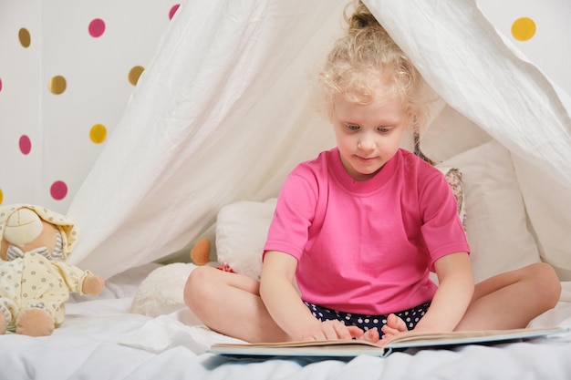 Una linda chica con el pelo rizado lee un libro en una cabaña de una sábana en la cama, una niña divirtiéndose y jugando en su tienda de campaña, Hut en la habitación de los niños