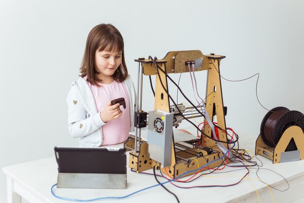 Linda chica con persianas impresas en 3D está mirando su impresora 3D mientras imprime su modelo 3D.