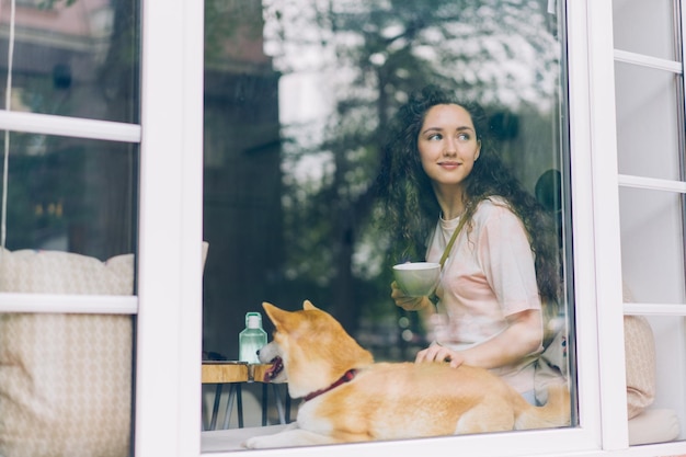 Linda chica relajándose en la cafetería con una taza de café acariciando a un lindo perro pedigrí