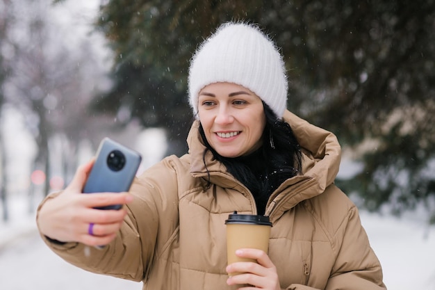 Linda chica riendo y haciendo una videollamada afuera en invierno Sostenga una taza de café desechable