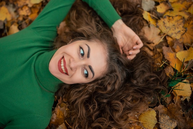 Linda chica rizada con los ojos abiertos acostados en las hojas de otoño sosteniendo la mano cerca de la cara y sonriendo Mujer mujer vistiendo suéter de cuello alto verde Retrato colorido de otoño Vista superior