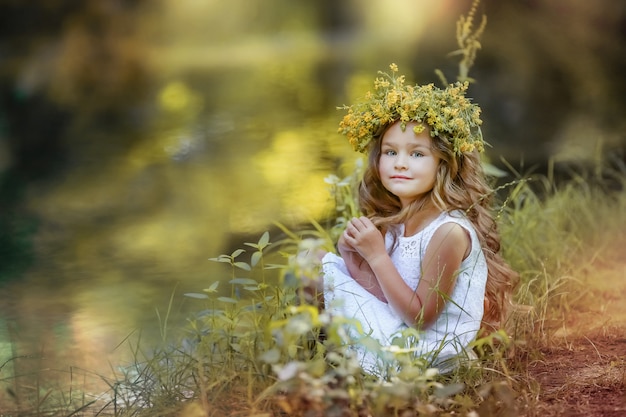 Linda chica rizada de pelo largo con una corona de flores silvestres amarillas en la cabeza se sienta en un césped verde