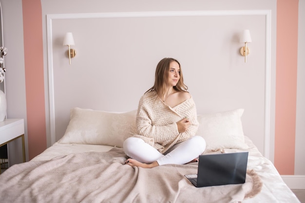 Una linda chica con ropa casera está sentada en una cama con una laptop