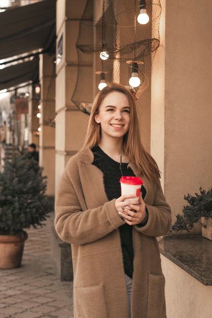 Linda chica en ropa de primavera, con un abrigo, de pie en la calle con una taza de café en sus manos, mirando hacia arriba y sonriendo contra el fondo de una pared de color beige