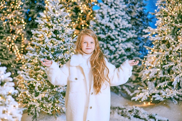 Linda chica rubia caucásica con abrigo de piel ecológica blanca caminando en el bosque de Navidad de invierno con luces.