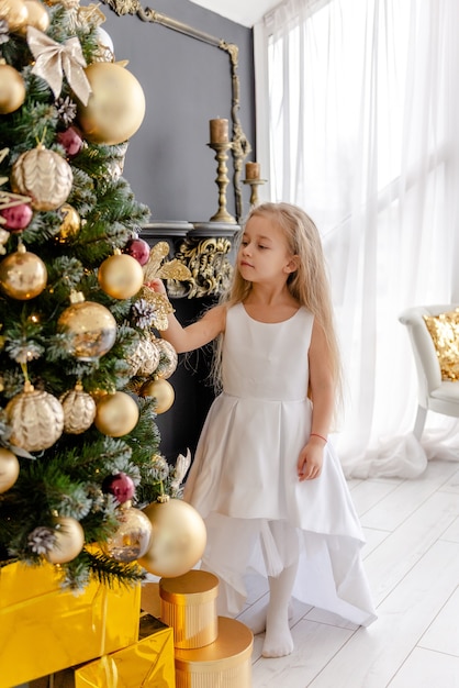 Una linda chica rubia decora un árbol antes de Navidad en casa. Infancia feliz. Decorar un árbol de Navidad antes de Navidad.