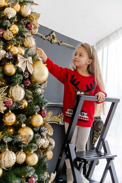 Una linda chica rubia decora un árbol antes de Navidad en casa. Infancia feliz. Decorar un árbol de Navidad antes de Navidad.