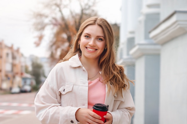 Linda chica rubia está caminando en la ciudad con una taza de café y sonriendo