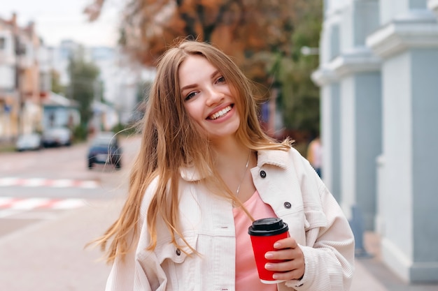 Linda chica rubia está caminando en la ciudad con una taza de café y sonriendo