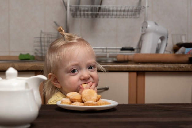 Linda chica rubia está desayunando en la cocina y mirando las galletas