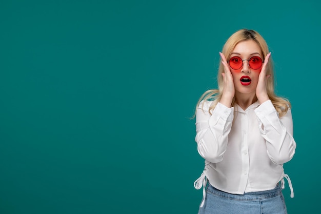 Linda chica rubia jovencita bonita en una camisa blanca con gafas de sol rojas sorprendida