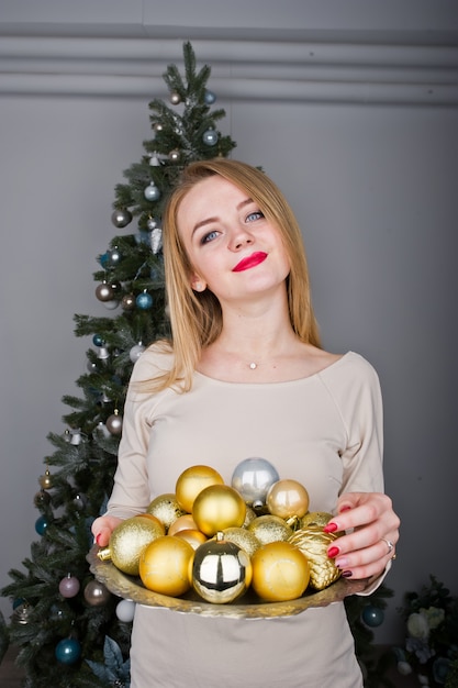 Linda chica rubia en vestido beige contra árbol de año nuevo con bolas doradas de año nuevo en el estudio