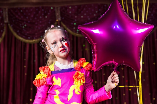 Linda chica rubia con vestido rosa festivo y maquillaje de payaso mientras sostiene un globo en forma de estrella, en un escenario