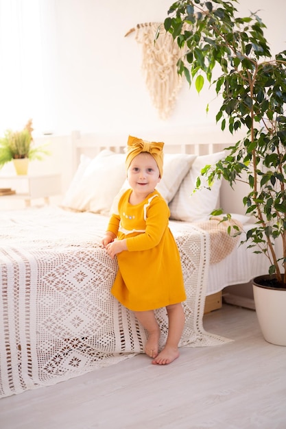 Una linda chica saludable con un vestido de algodón está parada sobre una alfombra en la luminosa sala de estar de la casa contra el fondo de la cama mirando a la cámara textiles para el hogar