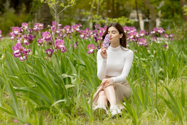 Linda chica sentada en un jardín floreciente sosteniendo lila oliendo con los ojos cerrados