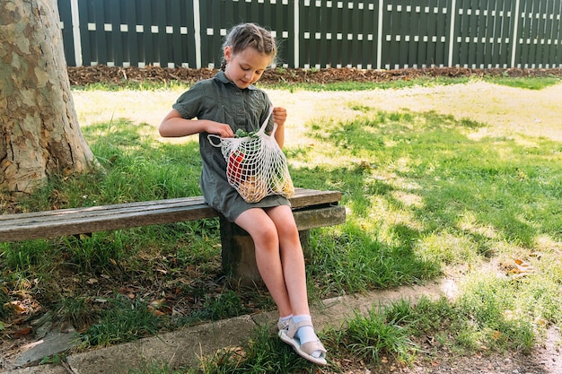 Una linda chica se sienta en un banco en la naturaleza con una bolsa de hilo de algodón con verduras
