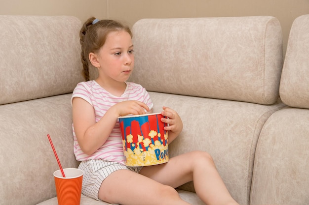 Linda chica se sienta en casa en el sofá viendo una película con entusiasmo comiendo palomitas de maíz y bebiendo cola