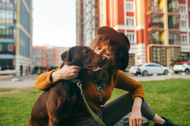 Linda chica con un sombrero se sienta en el césped con un perro marrón