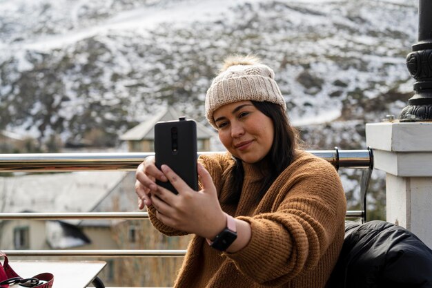 linda chica sonriendo tomando fotos mientras toma una copa en un pueblo nevado en las montañas