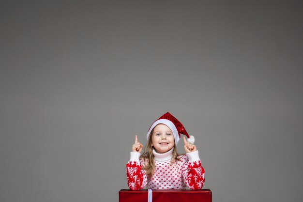 Linda chica sonriente con gorro de Papá Noel y suéter de invierno apoyándose en el regalo de Navidad con los dedos índices hacia arriba indicando que la idea vino a su mente. Copie el espacio.