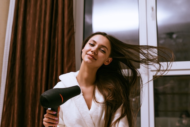 Linda chica sonriente se seca el pelo largo con un secador de pelo