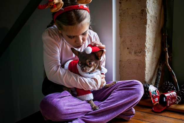 Linda chica sostiene al perro con sombrero rojo de santa y mira por la ventana esperando el milagro navideño de vacaciones