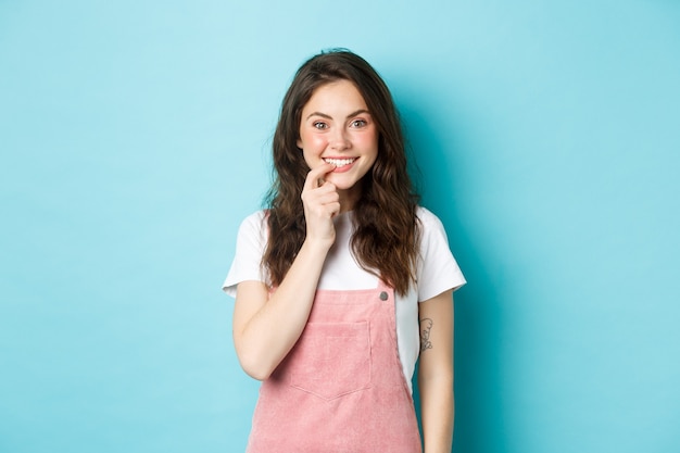 Linda chica tentada a probar o probar algo, mordiéndose el dedo y sonriendo, mirando con deseo e interés a la cámara, mirando el producto, de pie contra el fondo azul.