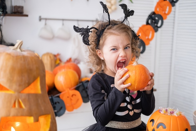 Linda chica en traje de bruja con calabaza en casa en la cocina, divirtiéndose, celebrando Halloween.