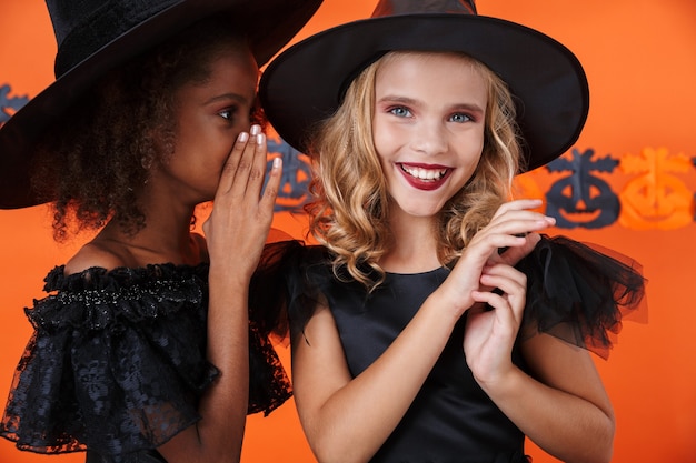 Linda chica en traje de halloween negro susurrando el secreto de un amigo sonriente en su oído aislado sobre la pared de calabaza naranja