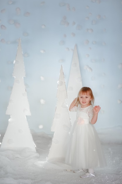 Linda chica en traje de princesa blanca entre árboles de Navidad brillantes artificiales blancos.