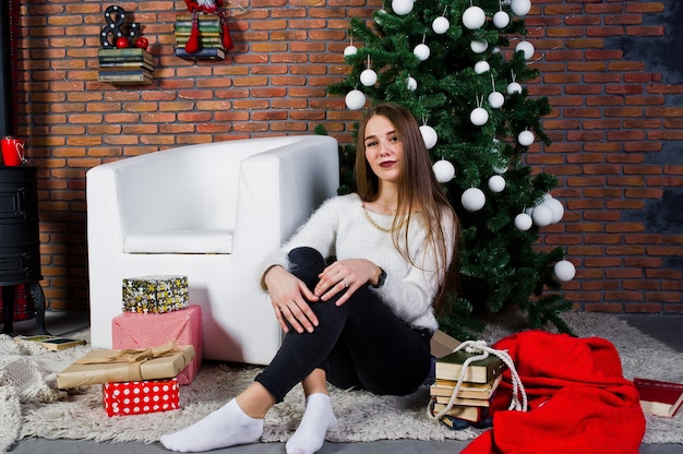 Linda chica usa suéteres calientes, pantalones negros contra el árbol de año nuevo con decoración navideña en el estudio.