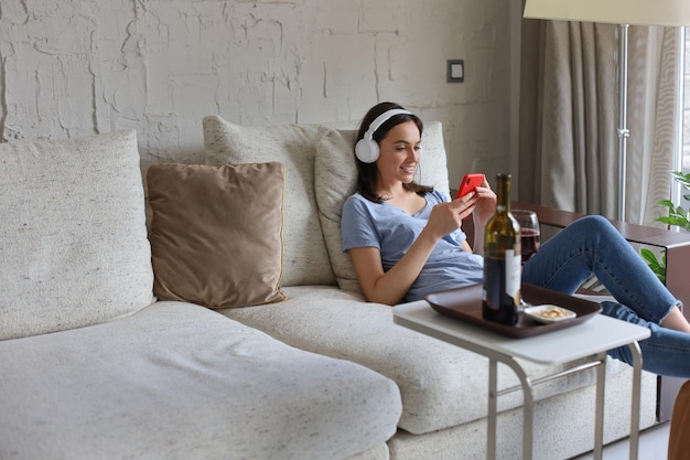 Linda chica usando su teléfono inteligente en el sofá en casa en la sala de estar Escuchando música bebiendo vino tinto relajación después de una dura semana en el trabajo