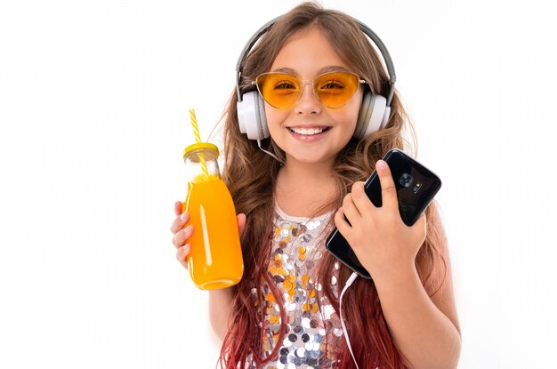 Linda chica en vestido brillante y gafas de sol de color amarillo brillante, con grandes auriculares blancos, escuchando música y sosteniendo un teléfono inteligente negro y una botella con jugo de naranja aislado