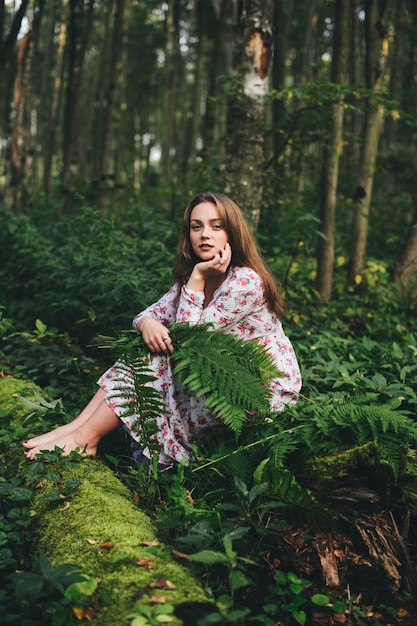 Una linda chica en un vestido de flores está sentada con un ramo de helechos en el bosque.