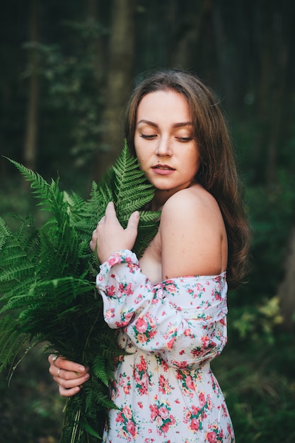 Una linda chica en un vestido de flores está sentada con un ramo de helechos en el bosque.