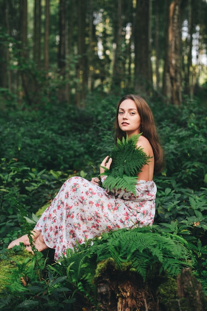 Una linda chica en un vestido de flores está sentada con un ramo de helechos en el bosque.