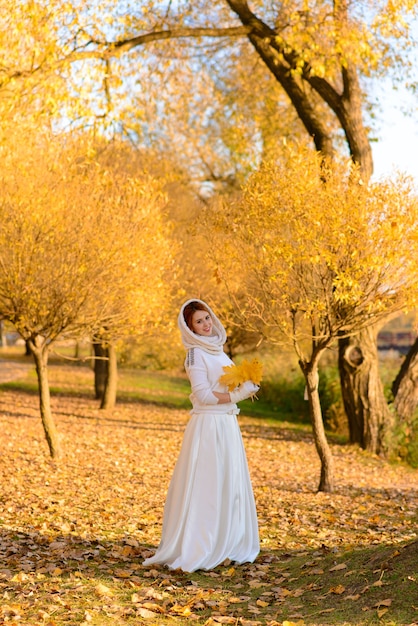 Linda chica en vestido de novia blanco largo posando en camino rural entre árboles otoñales en el bosque