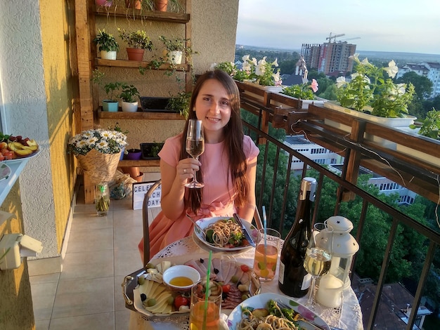 Linda chica en vestido rosa cena en el balcón. Mujer joven sentada y bebiendo limonada en la mesa con comida, bebidas y velas en la acogedora terraza con flores y luces en las noches de verano.