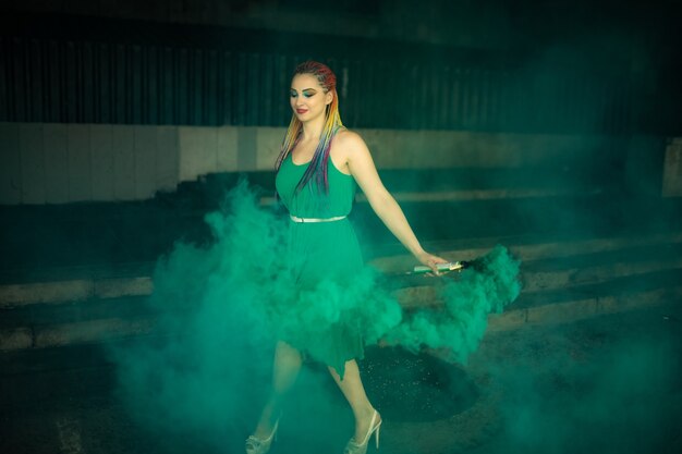 Linda chica con un vestido verde con trenzas africanas y maquillaje colorido posando con humo verde brillante en las calles de una ciudad de primavera cálida