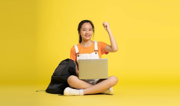 linda colegial asiática usando laptop e sentado sobre um fundo amarelo