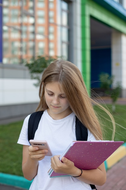 Linda colegial na rua com um smartphone, notebooks e uma mochila. Conceito de educação