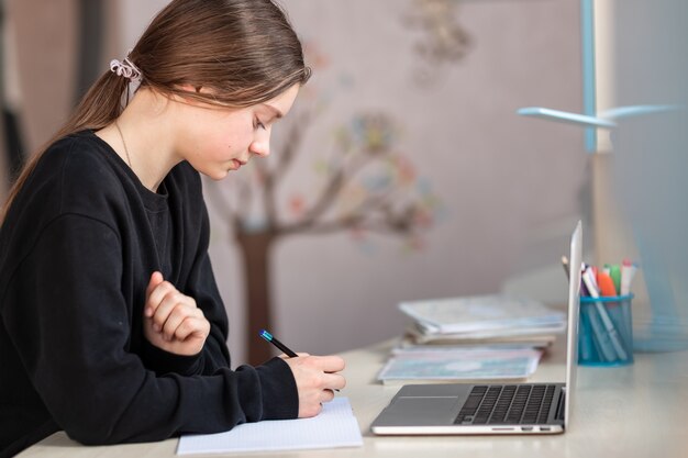 Linda colegial sorridente, estudando em casa, fazendo a lição de casa da escola. Livros de treinamento e caderno em cima da mesa. Educação a distância online